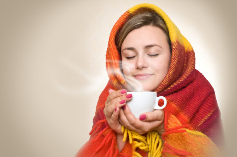 A young woman caught a cold, wrapped in a blanket and something hot drink from a cup.