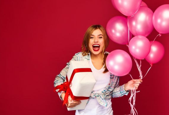 Excited young girl in hipster clothes. Woman model celebrating and holding box with gift present and pink air balloons on red background. St. Women's Day, Happy New Year, birthday holiday party